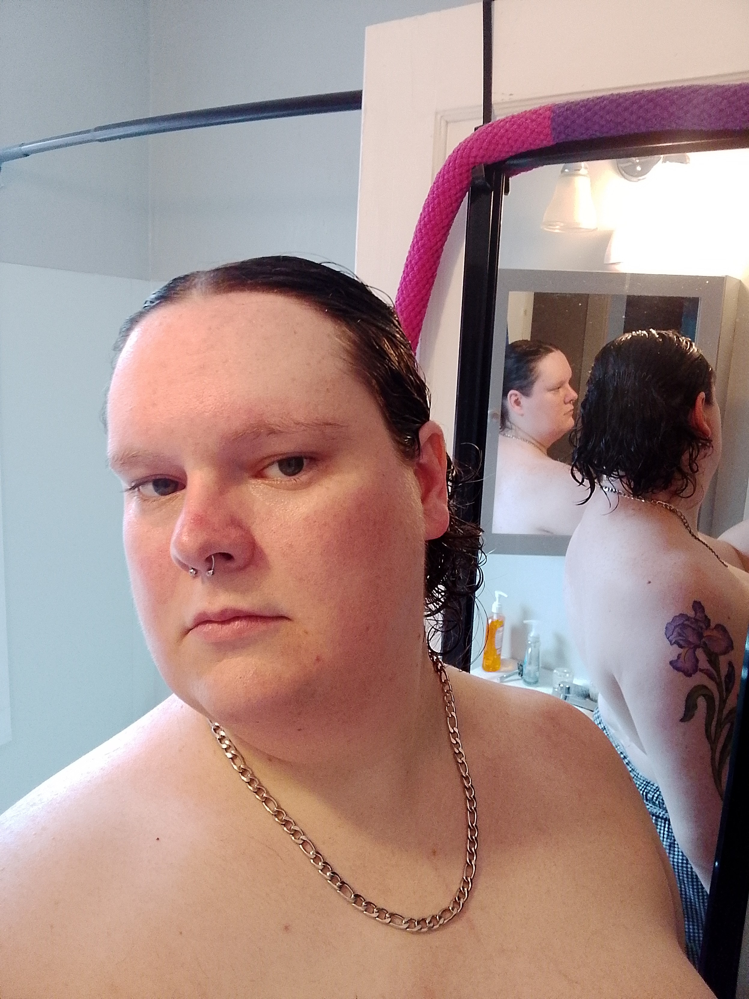 A photo of me, a plump white butch woman, shirtless wearing high-waisted pants and a silver chain, examining my wet pushed-back hair in two mirrors